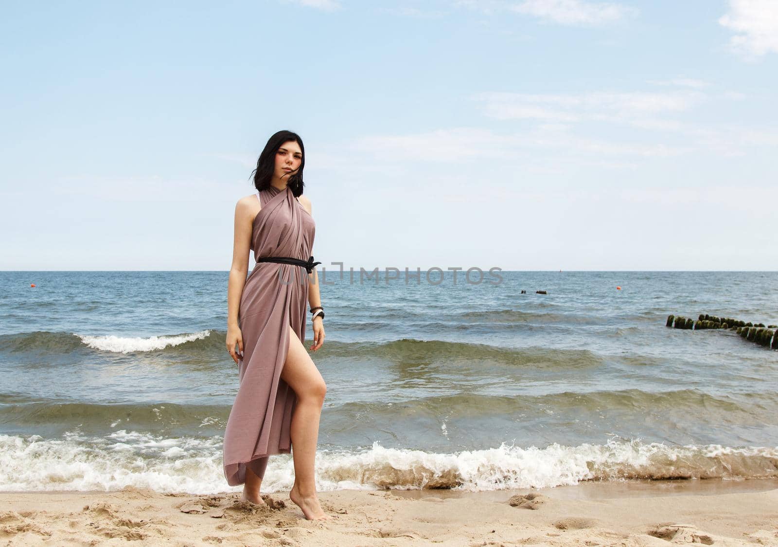 young brunette woman in beige dress stands on the sand by the sea on summer day