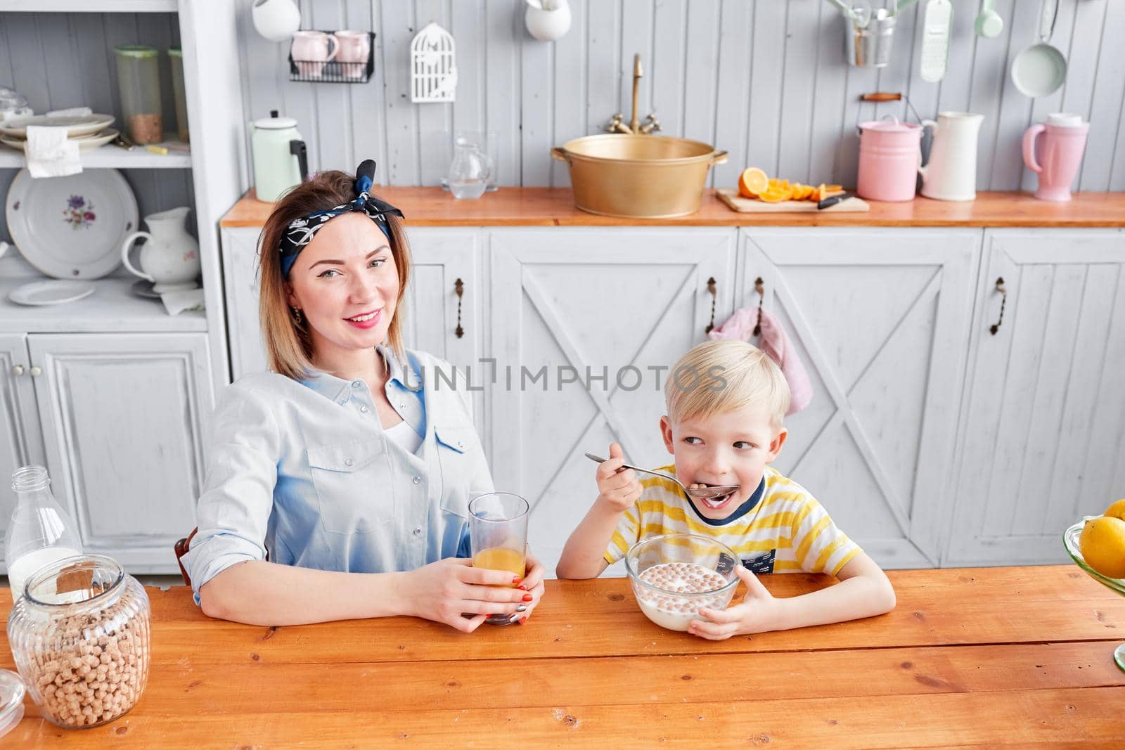 The boy greedily eats cereal with milk. Mother and son are smiling while having a breakfast in kitchen. Bright morning in the kitchen. Healthy Breakfast cereals and fresh fruit. by Malkovkosta