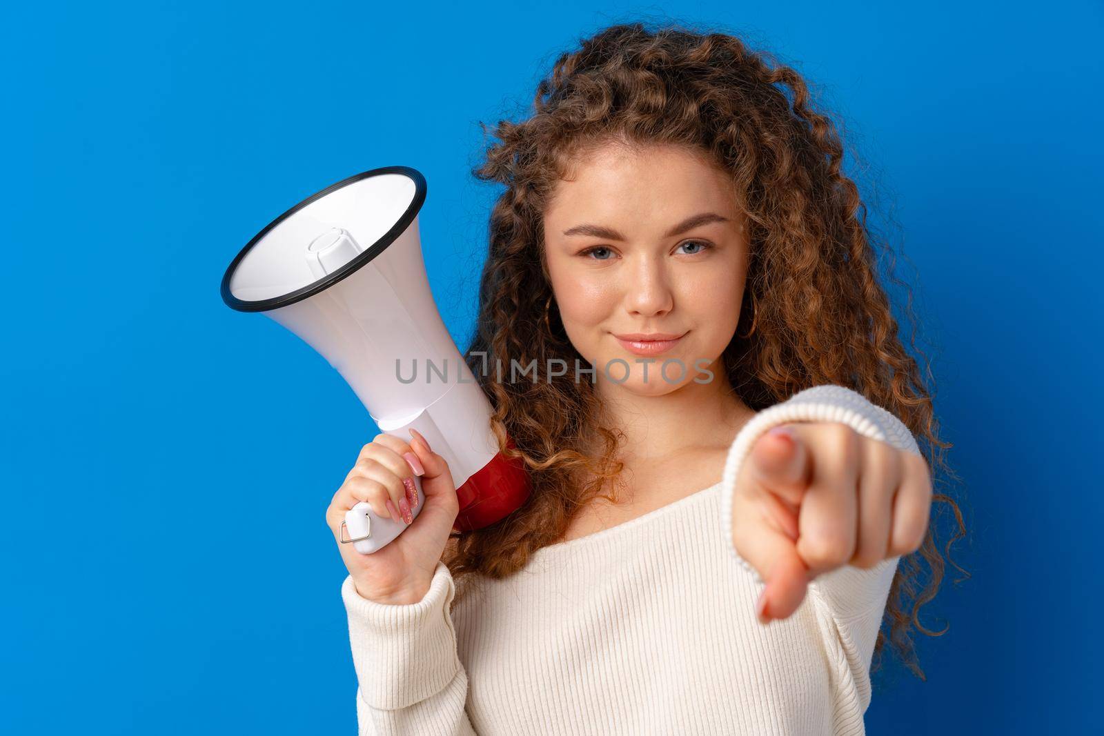 Young woman holding megaphone smiles and pointing to camera against blue background