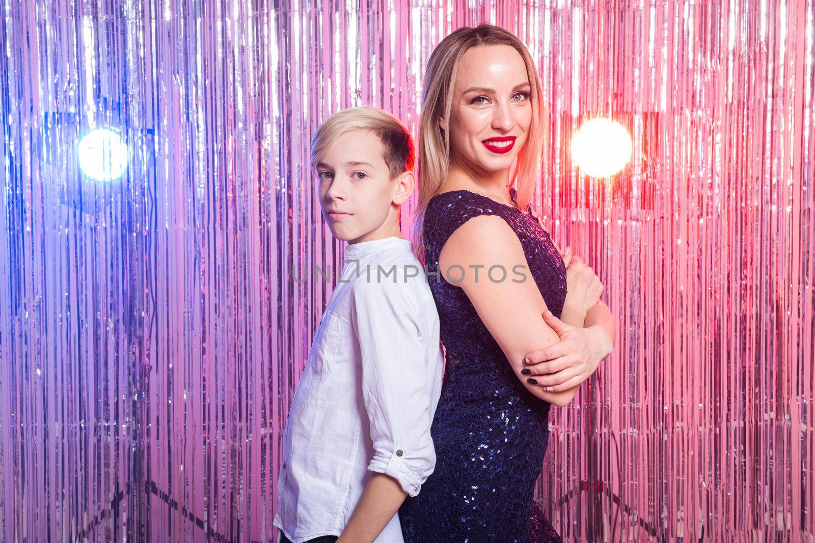 Mothers day, children and family concept - teen boy and his mom standing back to back on shiny party background