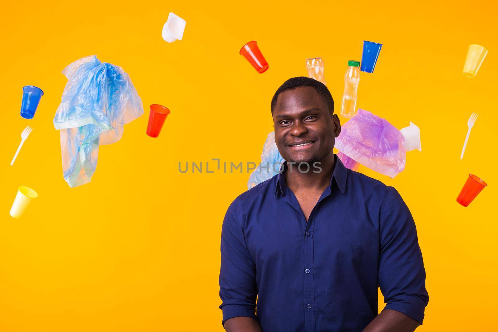 Problem of trash, plastic recycling, pollution and environmental concept - african american man is standing under trash on yellow background by Satura86