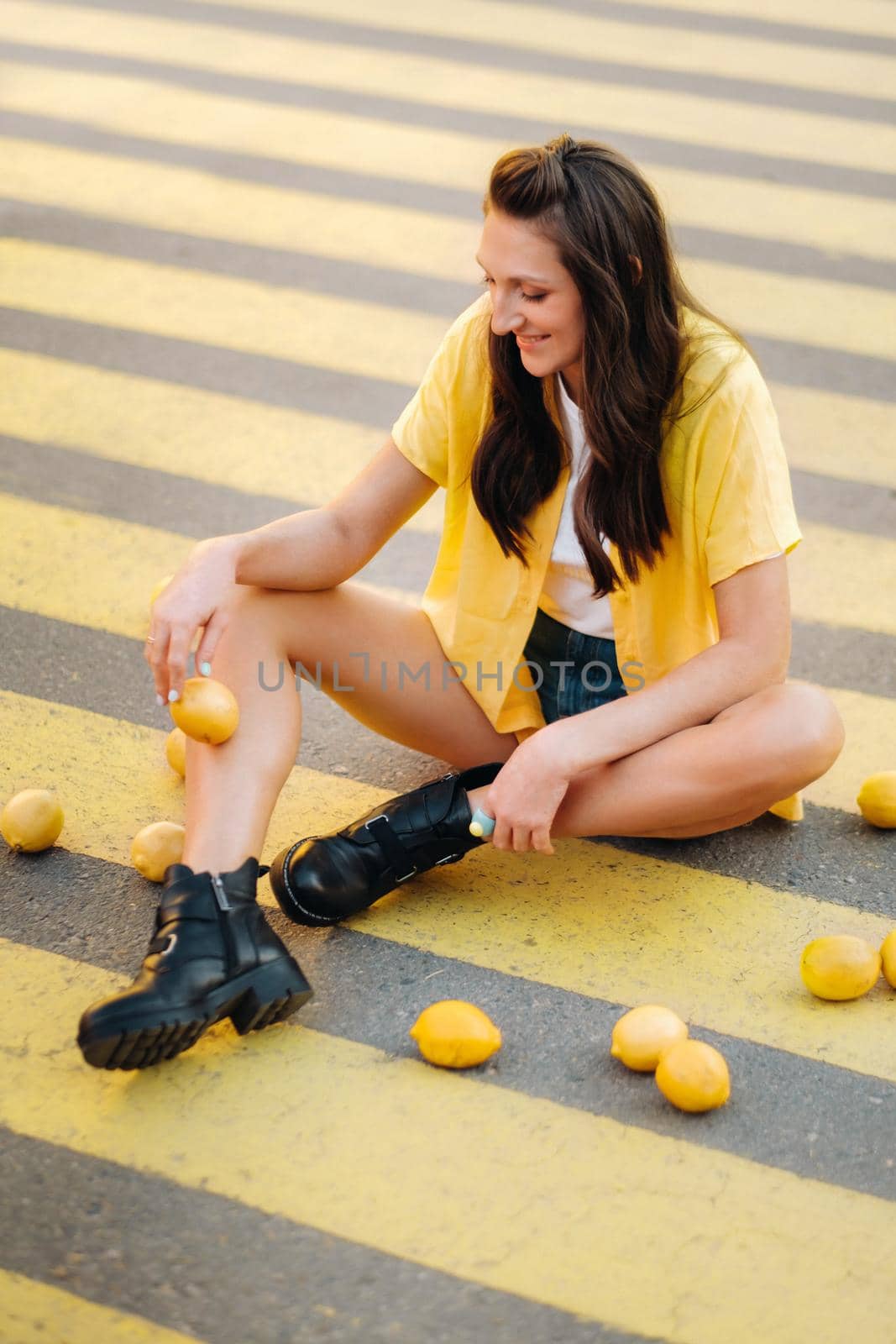 a girl with lemons in a yellow shirt, shorts and black shoes sits on a yellow pedestrian crossing in the city. The lemon mood.