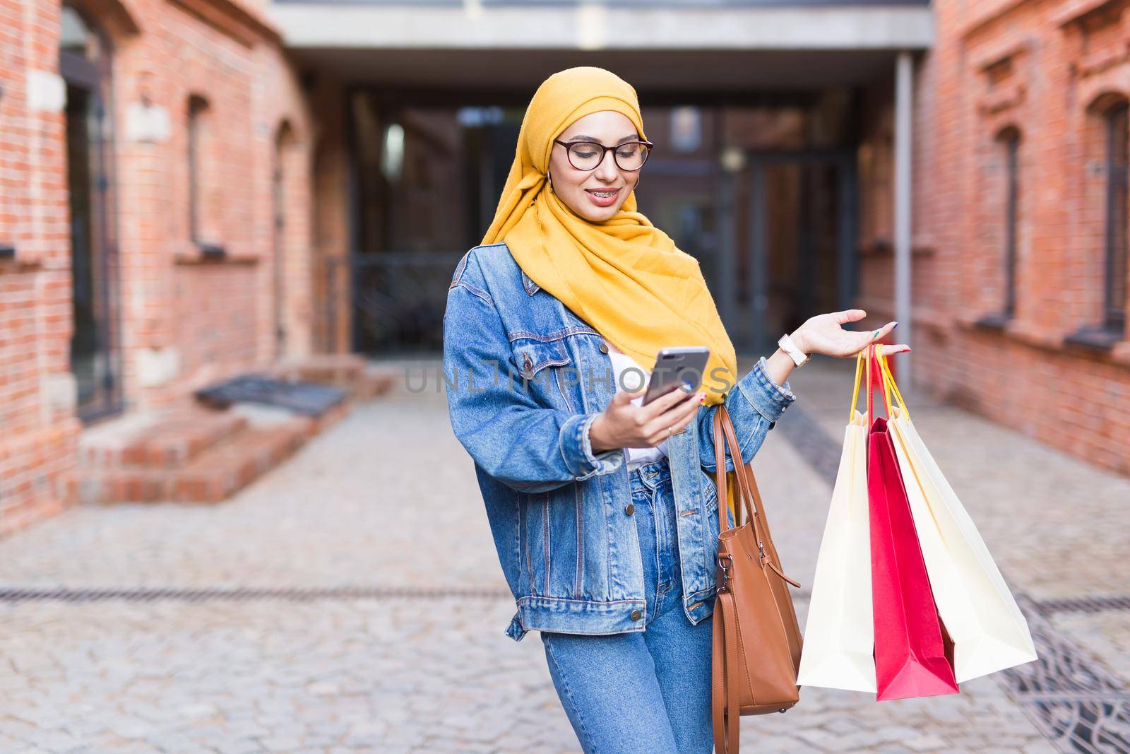 Sale and buying concept - Happy arab muslim girl with shopping bags after mall holds mobile phone by Satura86