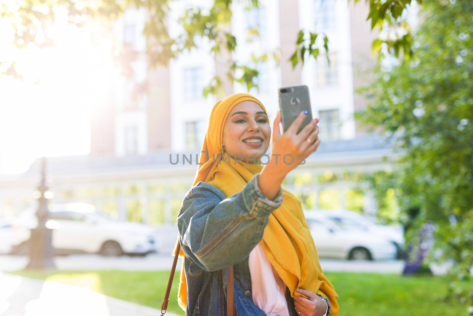 Muslim girl in hijab makes a selfie on the smartphone standing on the street of the city by Satura86