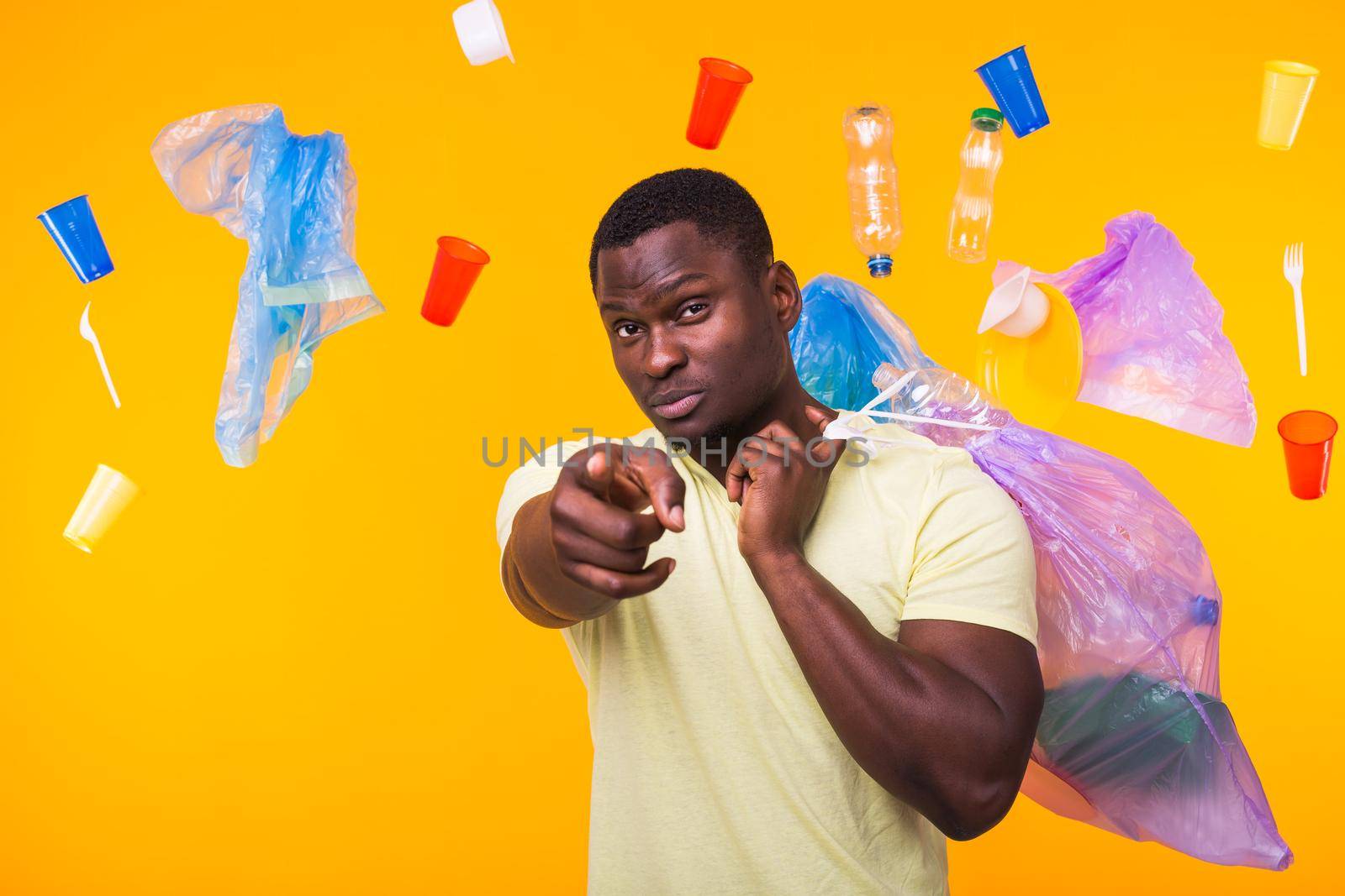Problem of trash, plastic recycling, pollution and environmental concept - funny african american man carrying garbage for recycling and pointing on you on yellow background. by Satura86