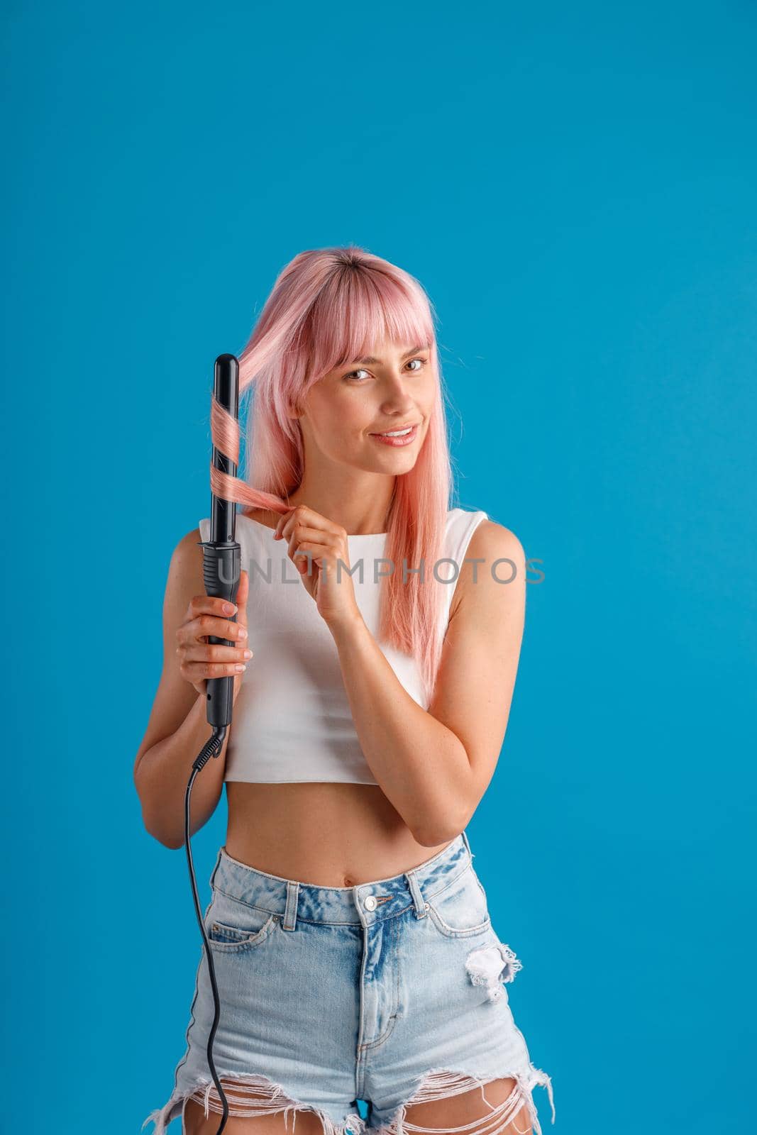 Cute woman smiling at camera while curling pink long straight hair using curling iron, standing isolated over blue studio background. Beauty and hair care concept