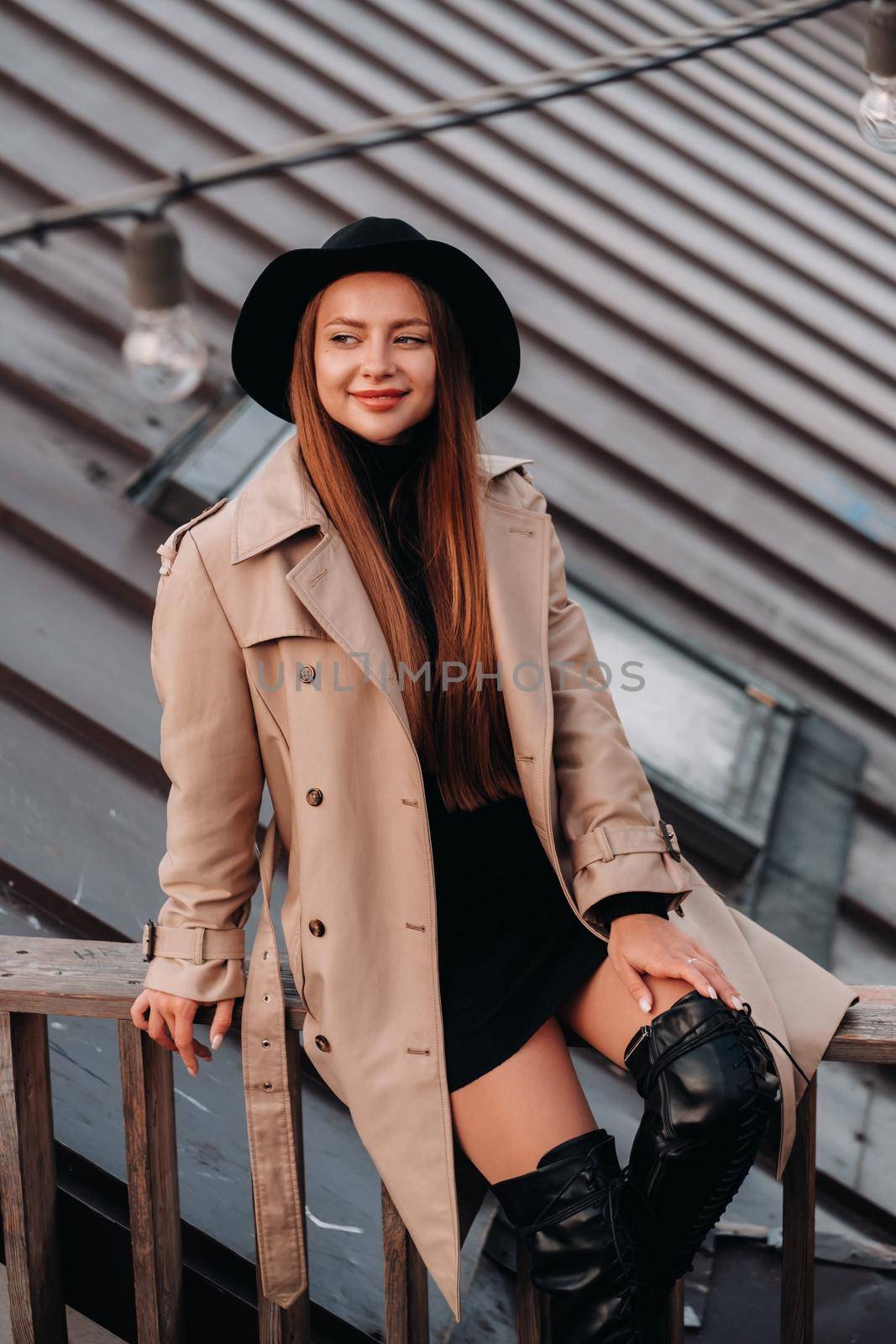 A stylish young woman in a beige coat and black hat sits on a rooftop in the city center. Women's street fashion. Autumn clothing.Urban style.