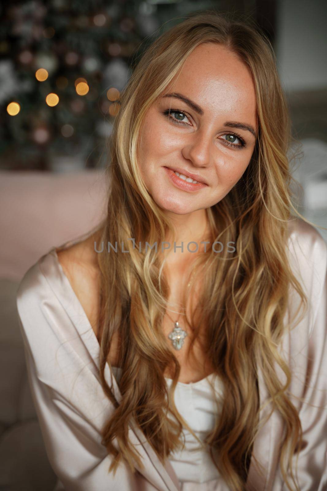 Portrait of a young blonde sitting on a sofa in pajamas. The room is decorated with a Christmas tree. by Matiunina