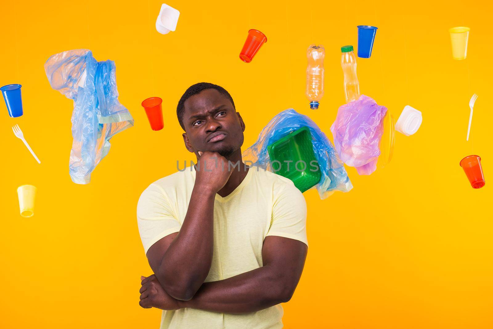Problem of trash, plastic recycling, pollution and environmental concept - Serious african american man looking on trash on yellow background. He is thinking about ecology by Satura86