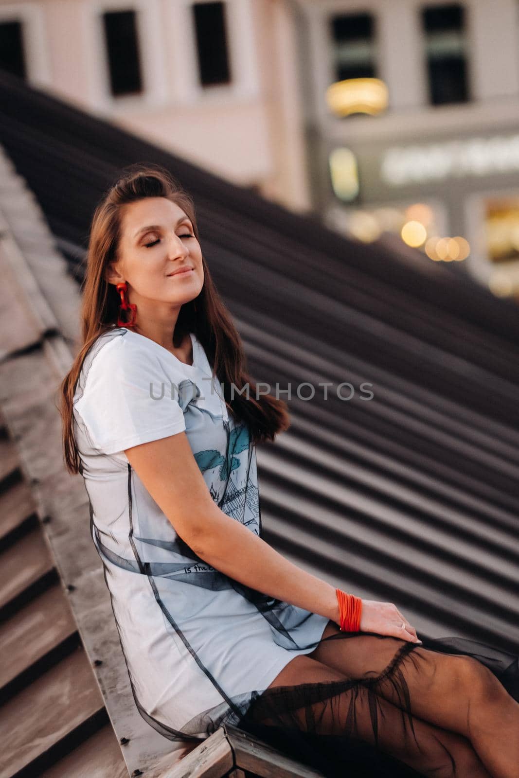 A girl sits on the roof of a house in the city in the evening. Portrait of a model in a dress and sneakers