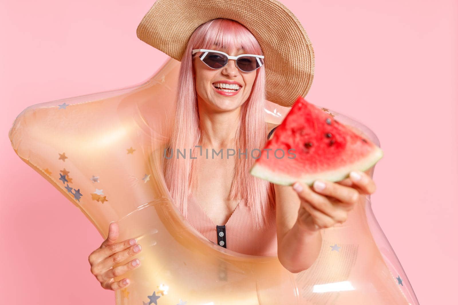 Excited woman with pink hair in swimsuit and sunglasses holding watermelon slice and inflatable star for swimming isolated over pink background. Summer vacation, fun, party concept