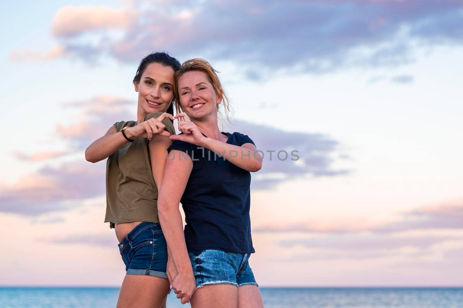 leisure and friendship concept - two happy smiling teenage girls or best friends at seaside making hand heart gesture at sunset.