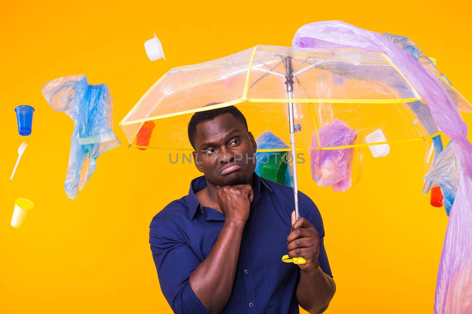 Problem of trash, plastic recycling, pollution and environmental concept - Serious african american man looking on trash on yellow background. He is thinking about ecology.