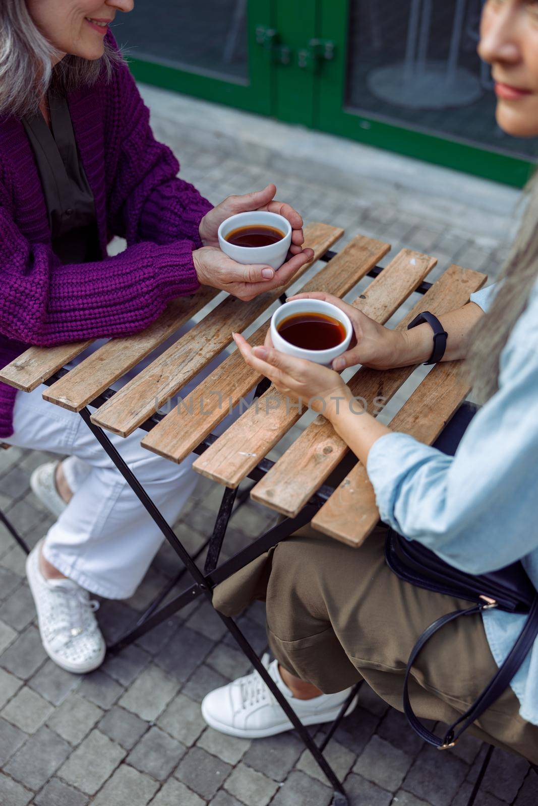 Senior ladies old friends hold cups of delicious coffee sitting at small table on outdoors cafe terrace on autumn day closeup