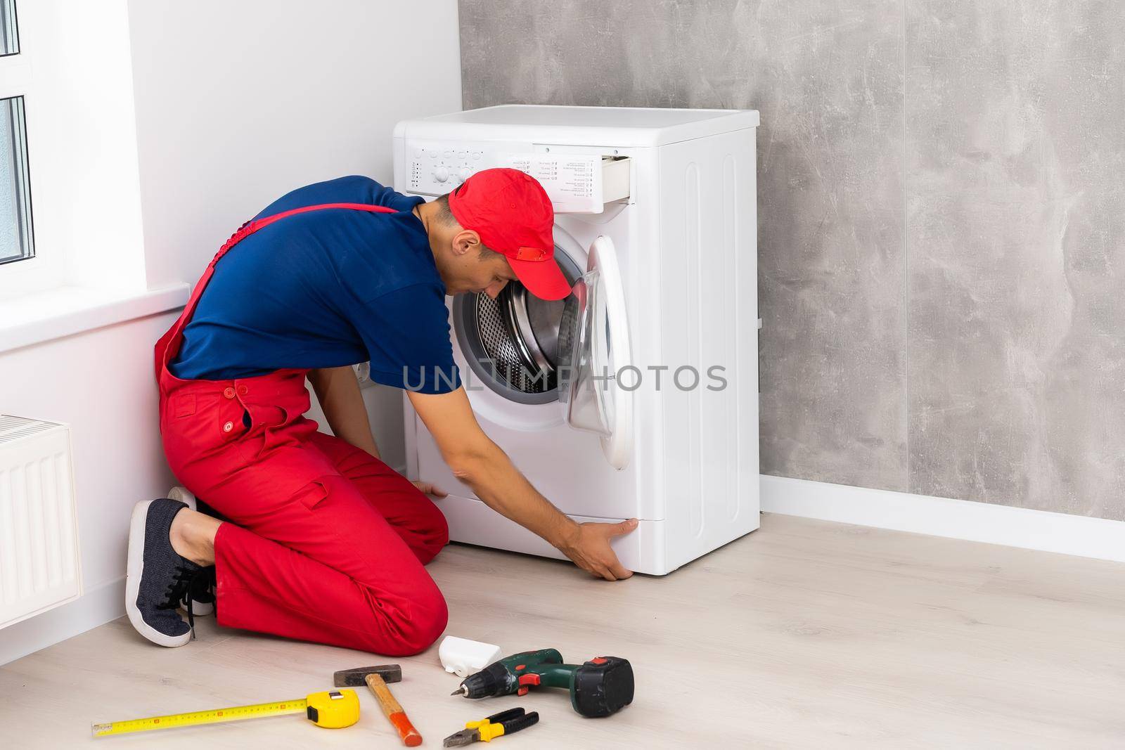 Plumber in overalls with tools is repairing a washing machine in the house