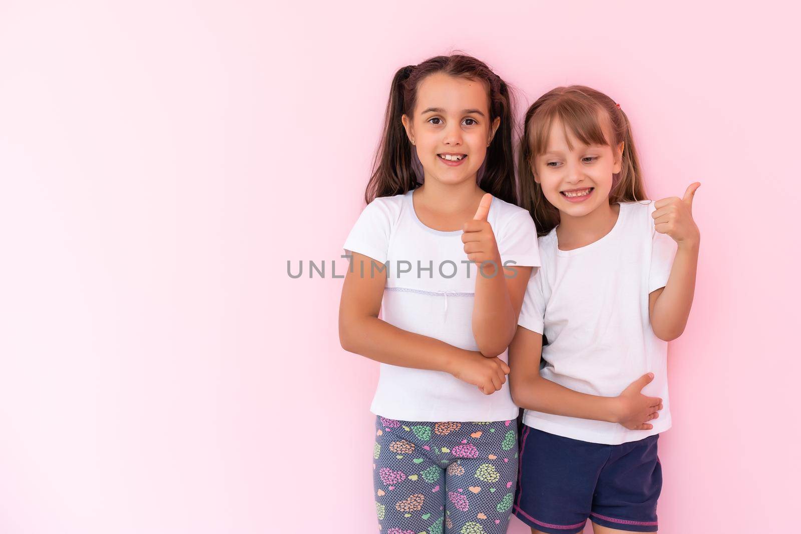 Children showing like. Two adorable happy little girls gesturing thumbs up together and smiling to camera, excellent feedback, good job. indoor studio shot isolated on pink background