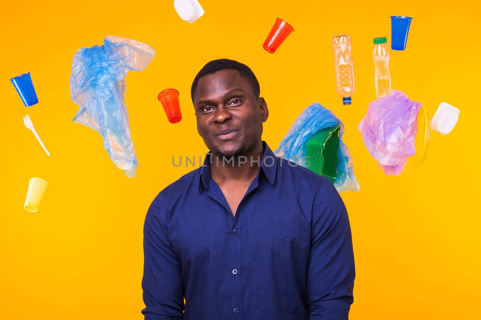 Problem of trash, plastic recycling, pollution and environmental concept - african american man is standing under trash on yellow background.