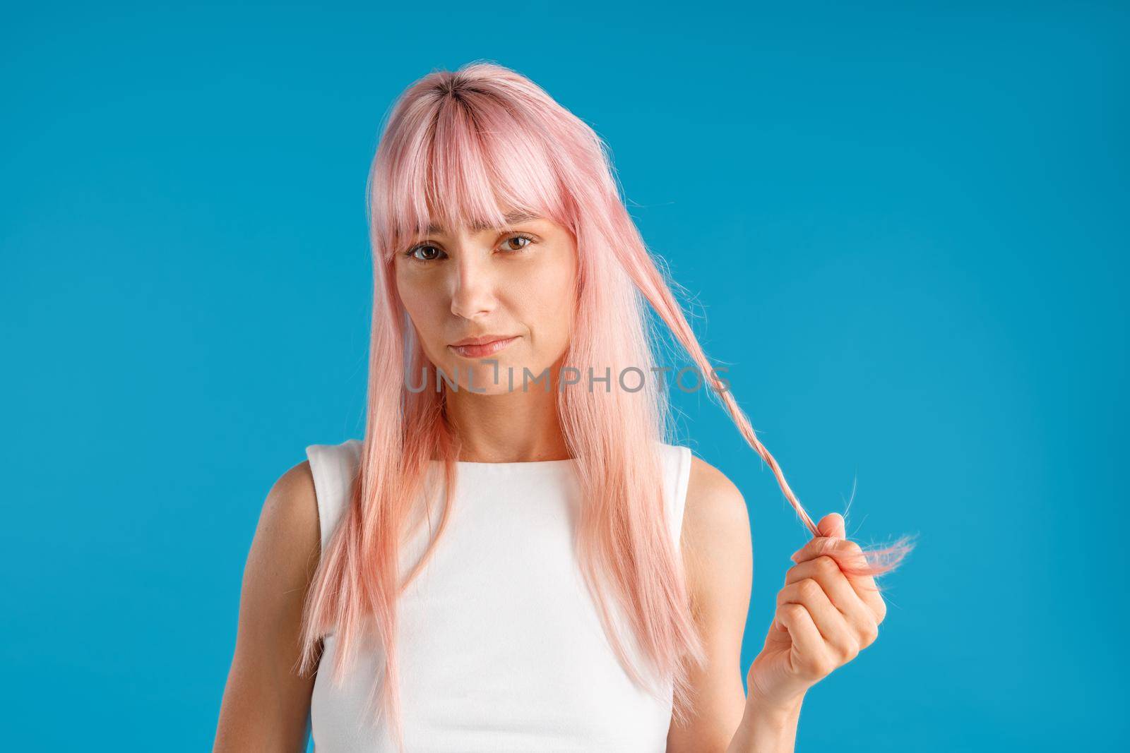 Young woman with natural long pink dyed hair holding a strand of it and looking upset at camera, posing isolated over blue studio background by Yaroslav_astakhov