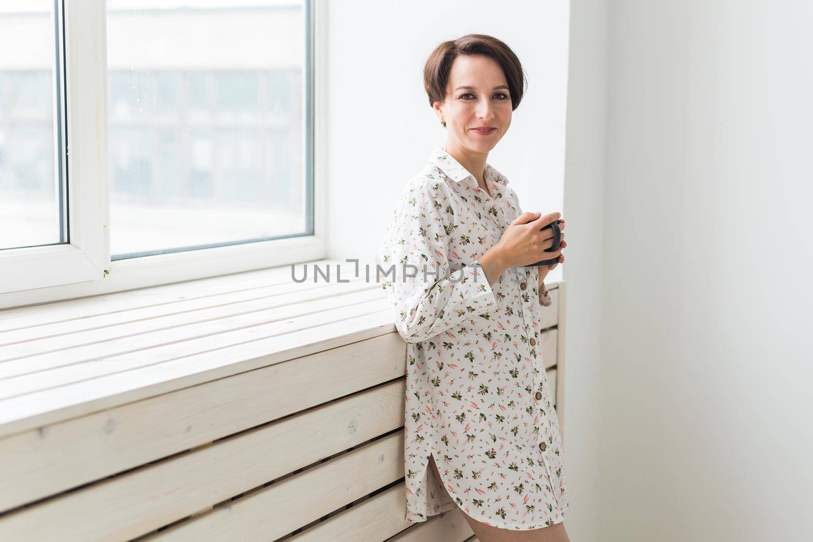 Beautiful young woman with cup of tea standing near the window at home