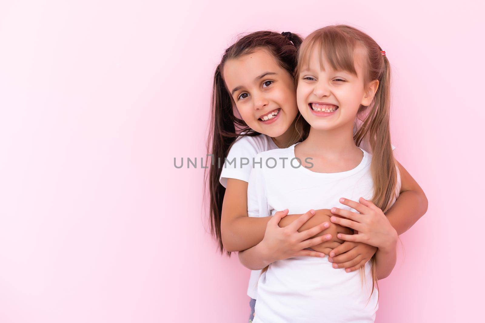 Two little girls hugging each other. Isolated on on a pink background