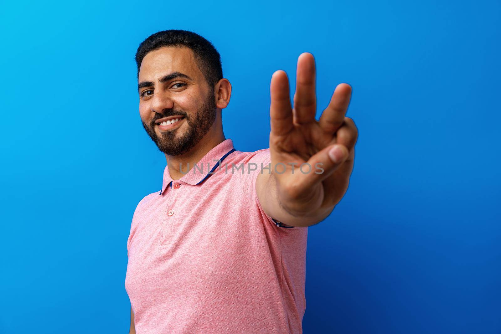 Young arabian man smiling and showing three fingers against blue background, close up