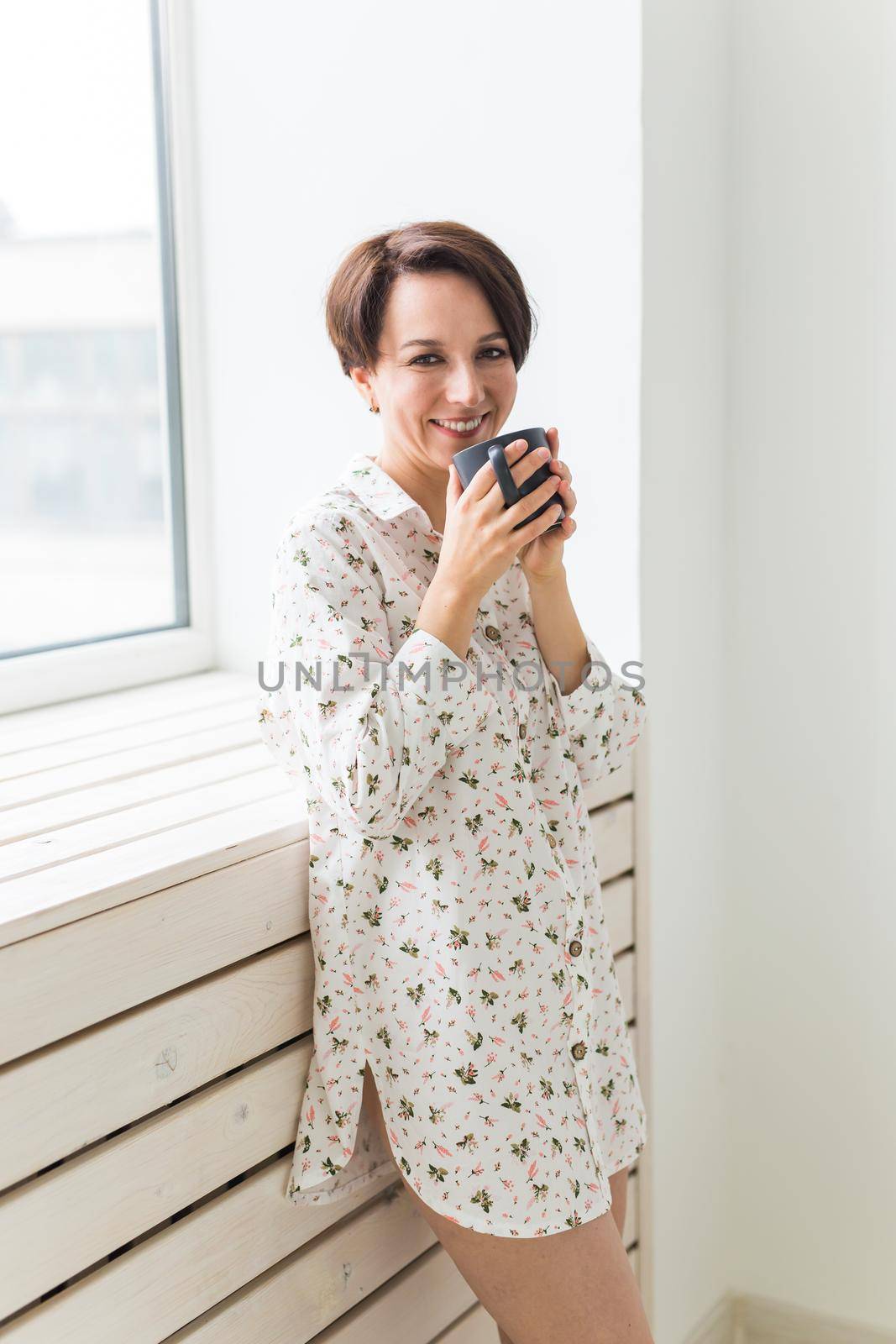 Calm and coziness. Beautiful young woman with cup of tea standing near the window at home by Satura86