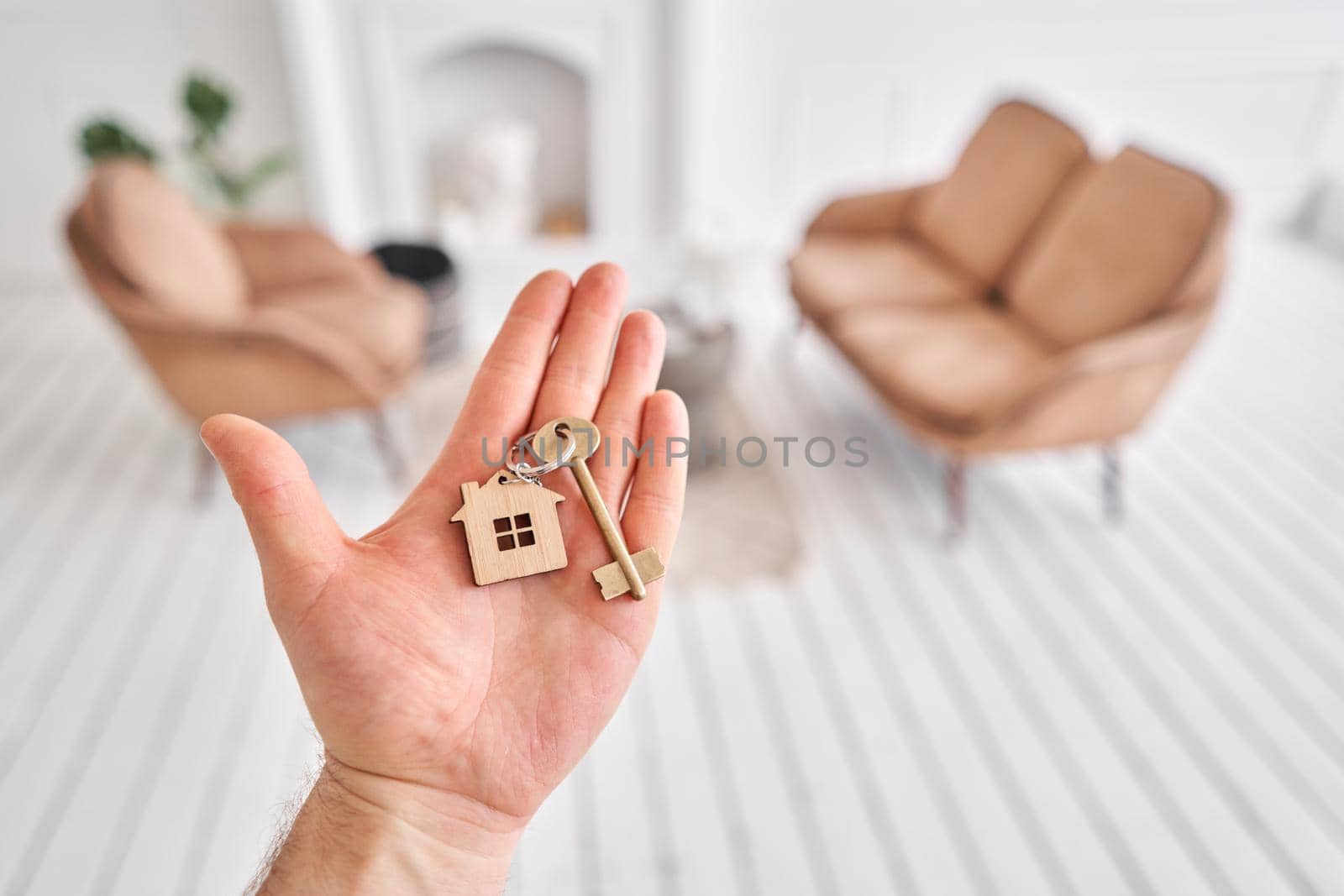 Men hand holding key with house shaped keychain. Modern light lobby interior. Mortgage concept. Real estate, moving home or renting property. by Malkovkosta
