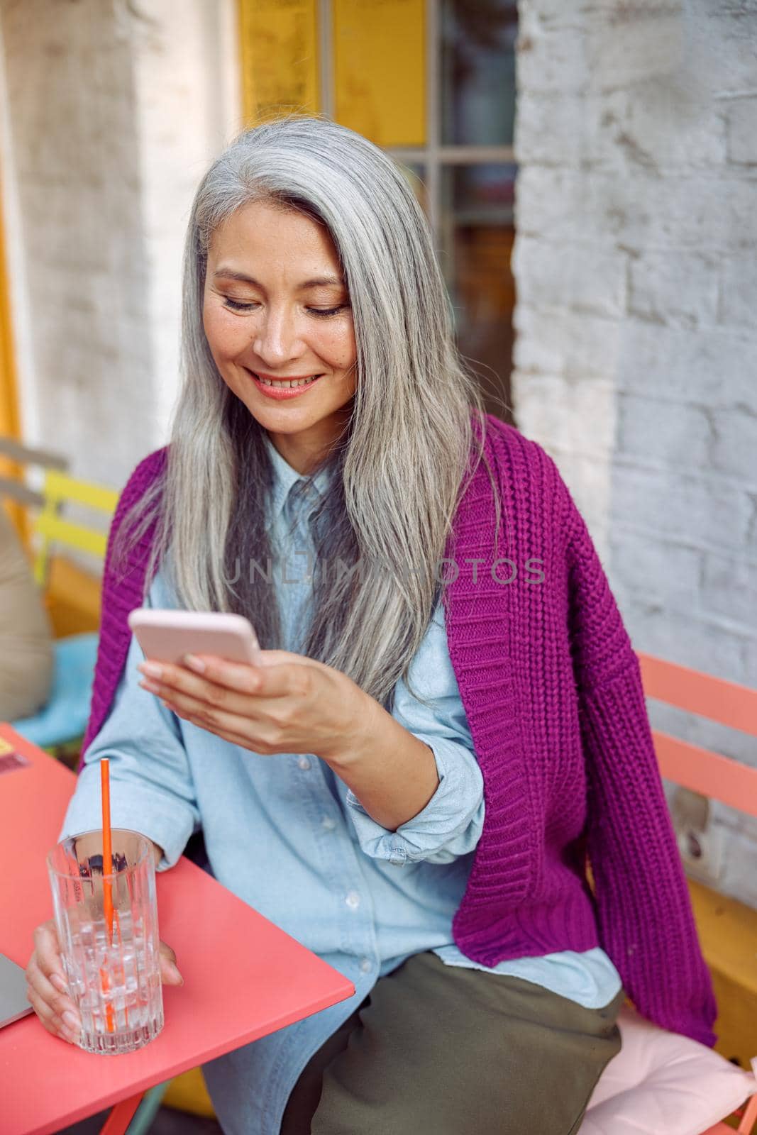 Pretty senior Asian lady with grey hair reads sms on mobile phone holding drink at table on outdoors cafe terrace