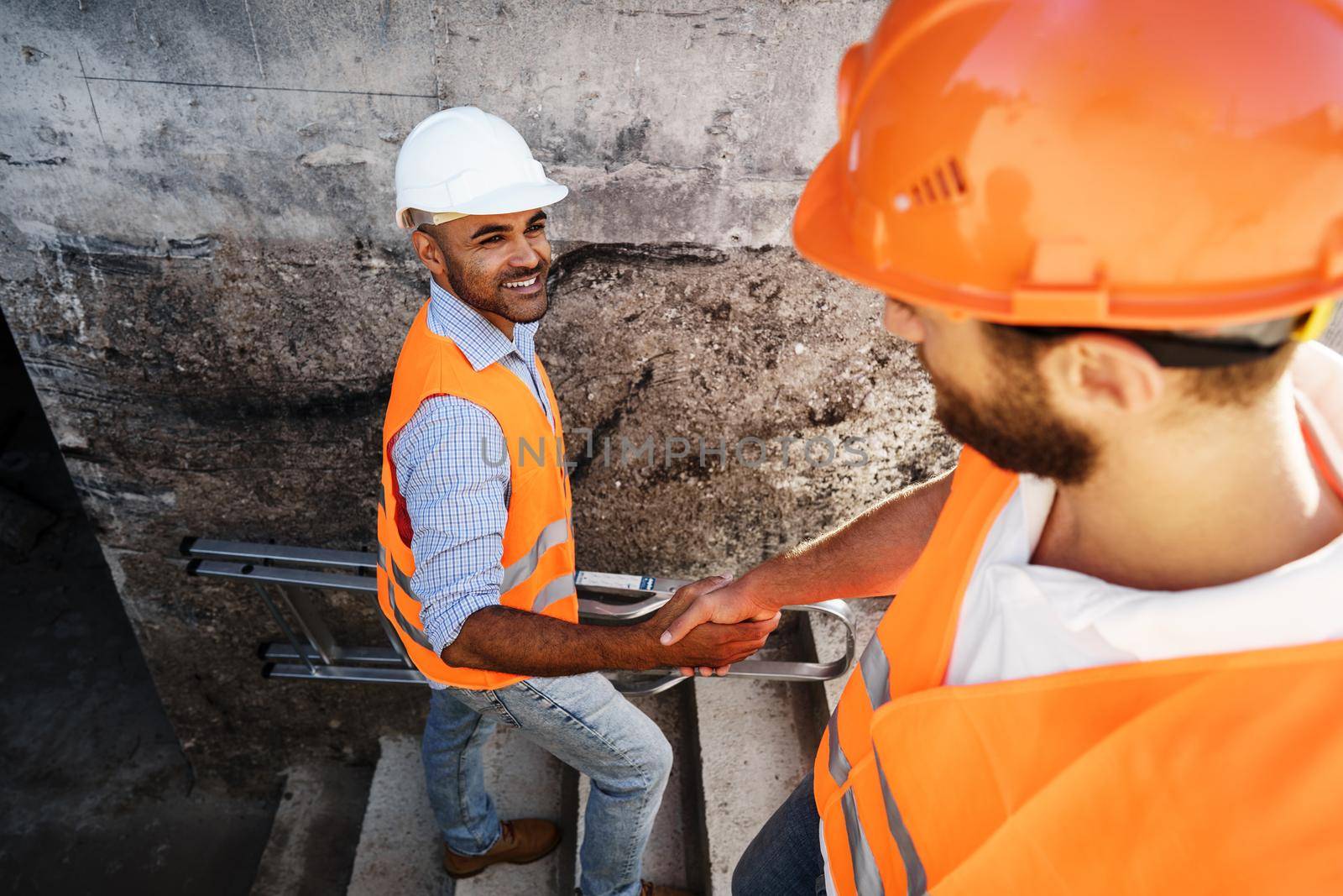 Two men engineers in workwear shaking hands against construction site, close up