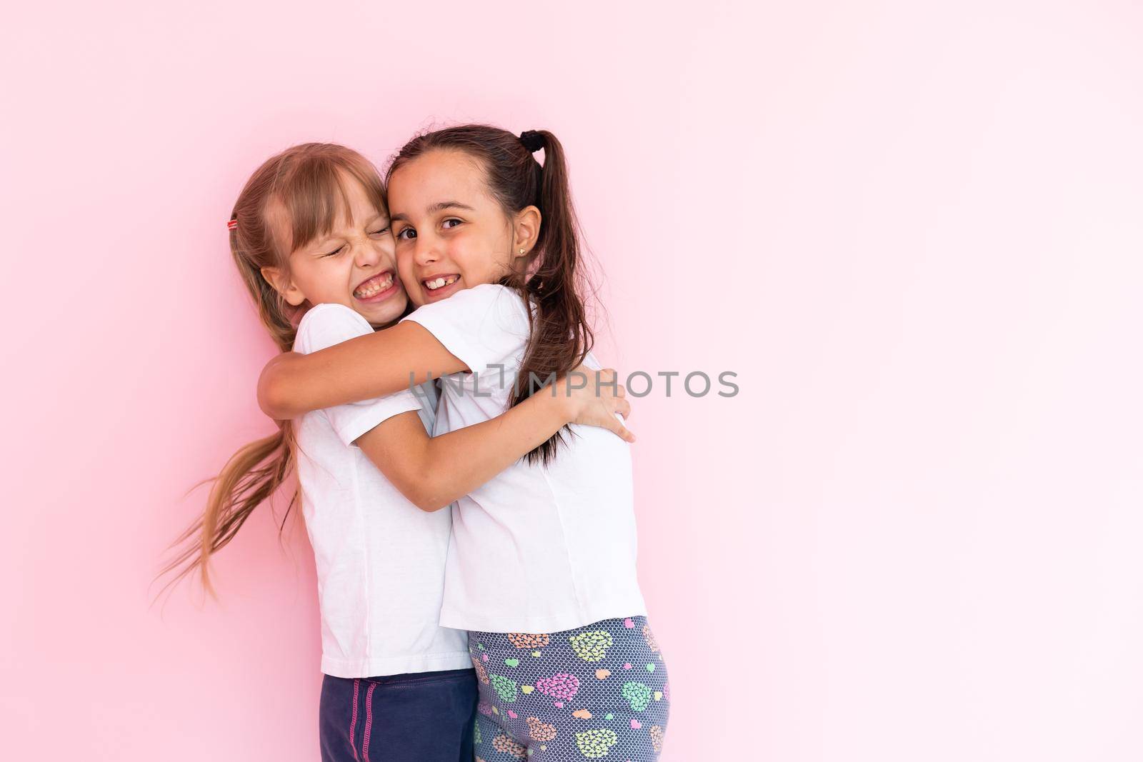 Two little girls hugging each other. Isolated on on a pink background