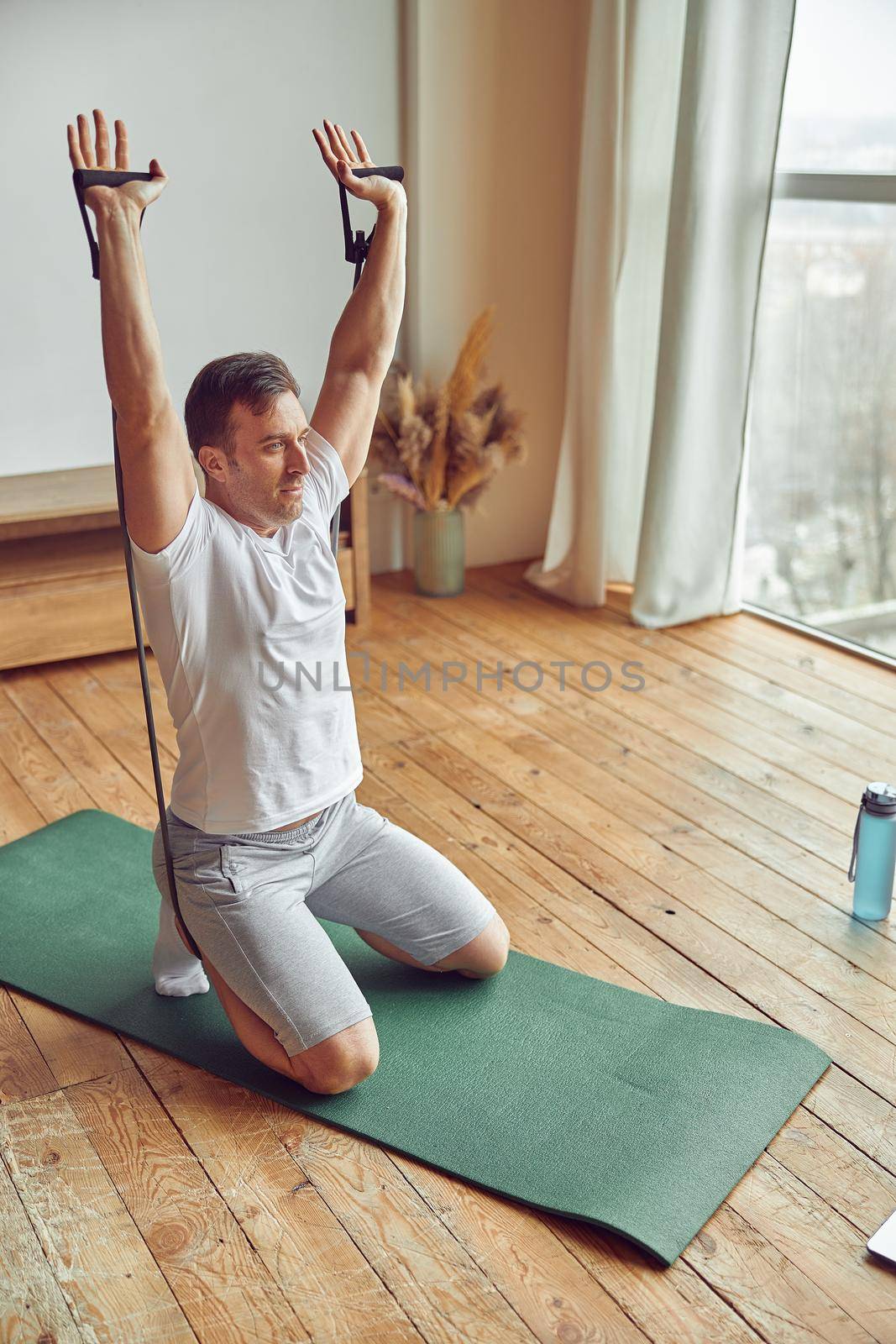 Top view of motivated male sitting on mat and and lifting arms with resistance bands while watching laptop