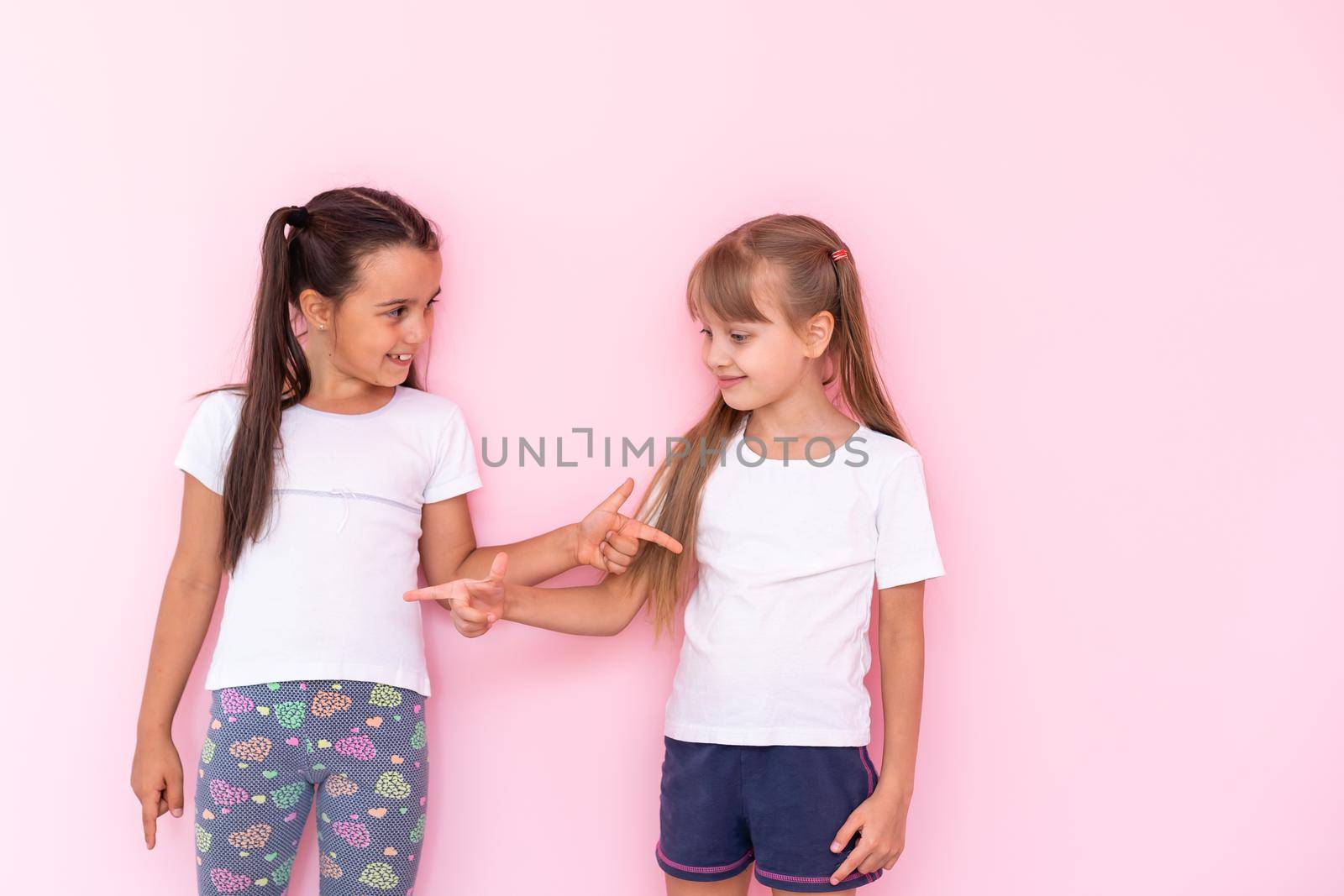 Amazed little kids girls showing white t-shirt, isolated on pink background. Childhood lifestyle concept. Mock up copy space. Pointing index finger aside by Andelov13
