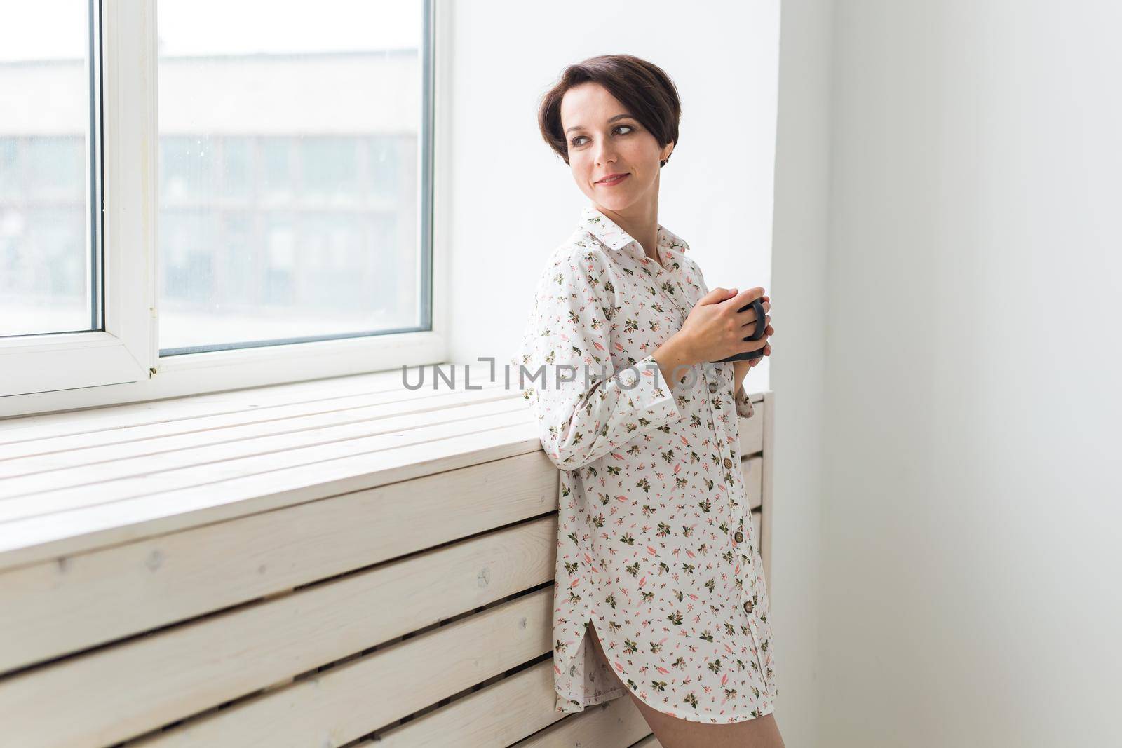 Beautiful young woman with cup of tea standing near the window at home