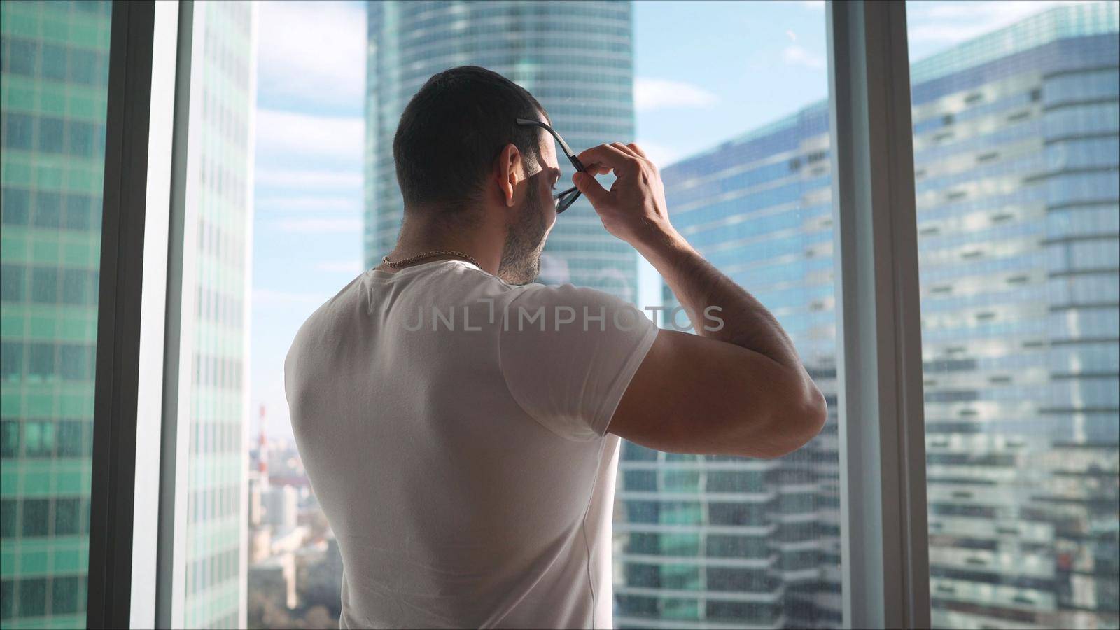 Serious tough young man in sunglasses with arms crossed. A guy in sportswear is standing in sunglasses against the background of panoramic windows.