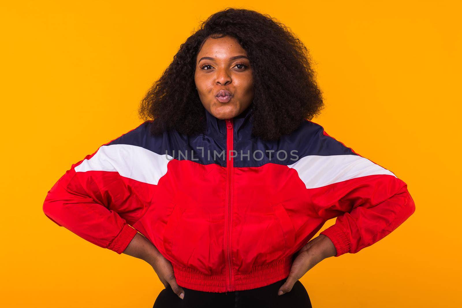 Funny curly african american girl dressed in the red sports jacket in the studio on yellow background. Fashion and people.