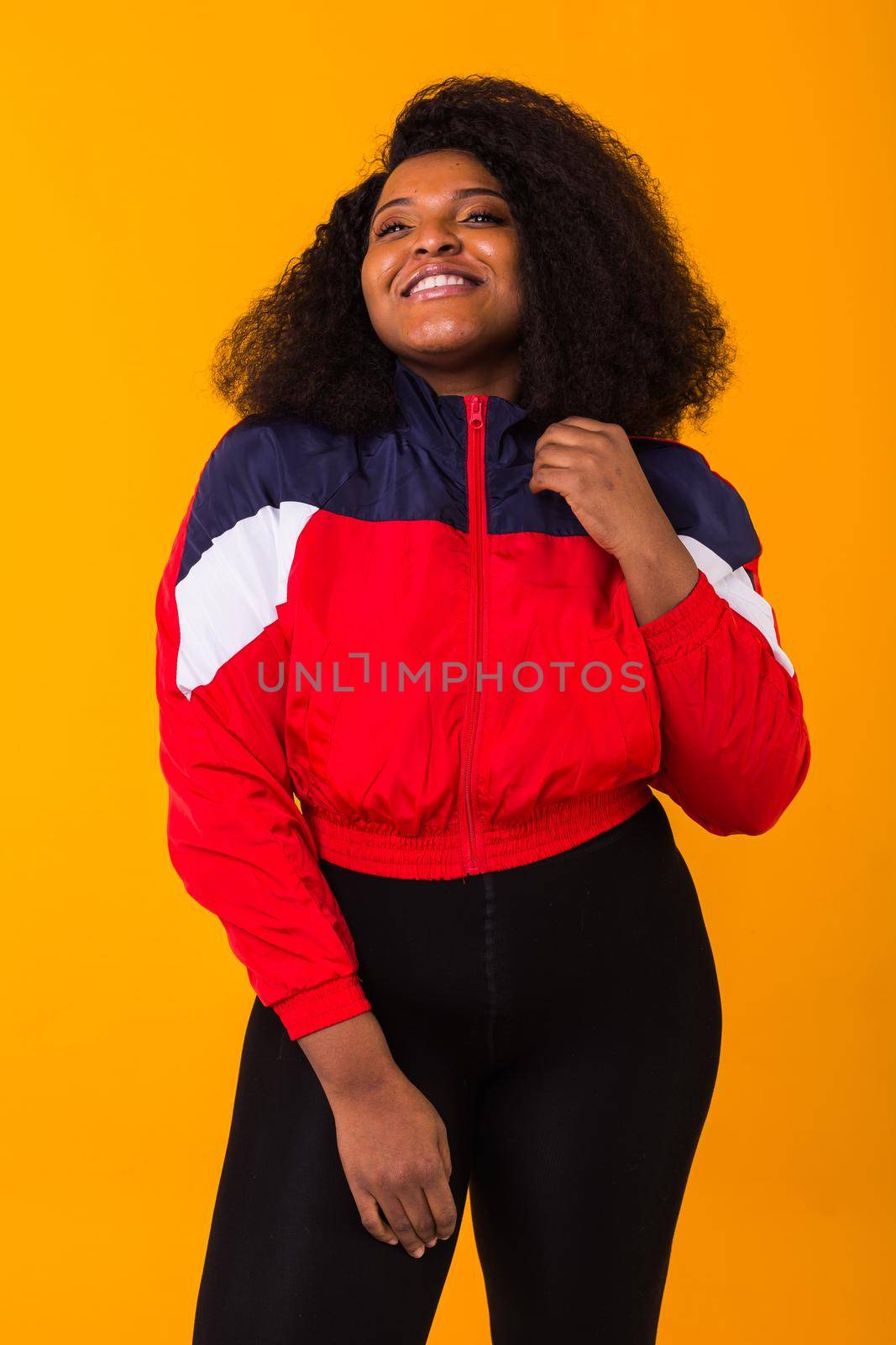 Funny curly african american girl dressed in the red sports jacket in the studio on yellow background. Fashion and people.