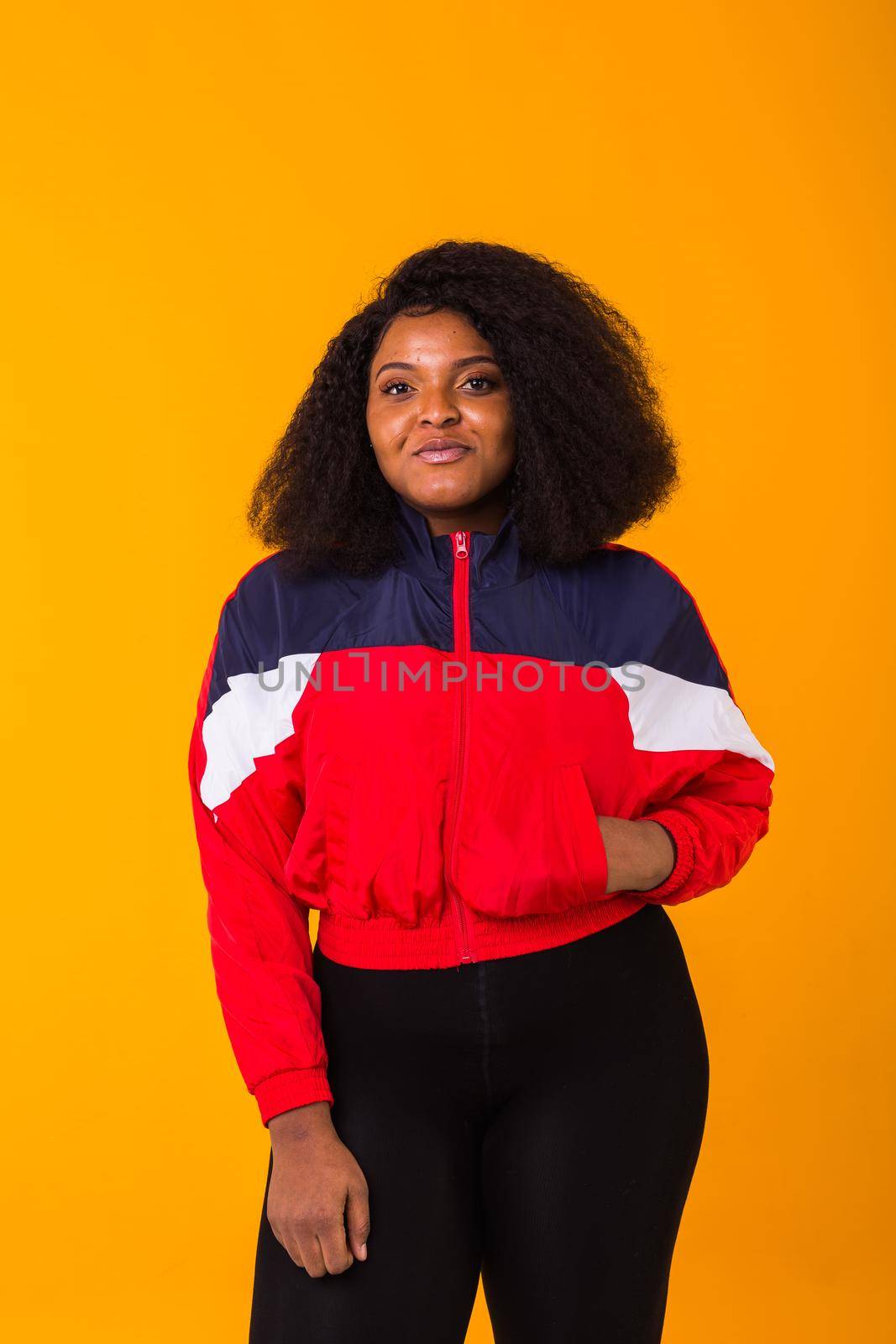 Funny curly african american girl dressed in the red sports jacket in the studio on yellow background. Fashion and people.