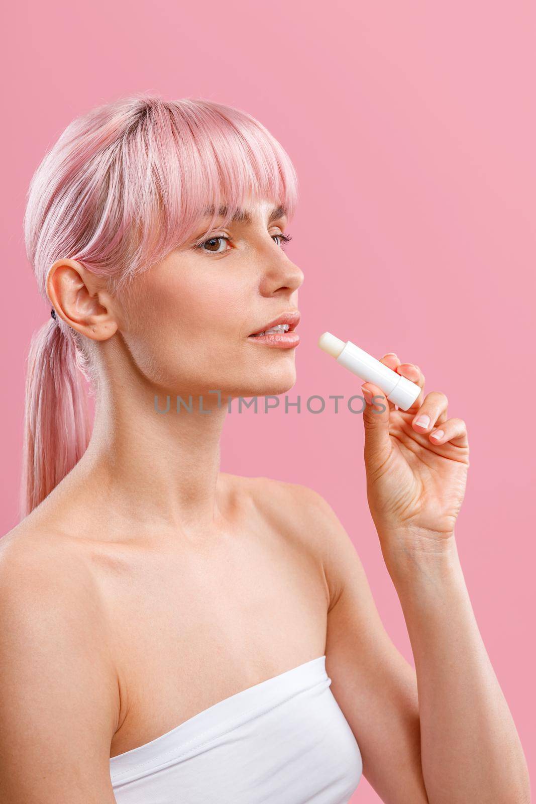 Beautiful young woman with pink hair and naked shoulders holding lip balm near her lips isolated over pink background. Beauty, lip care concept