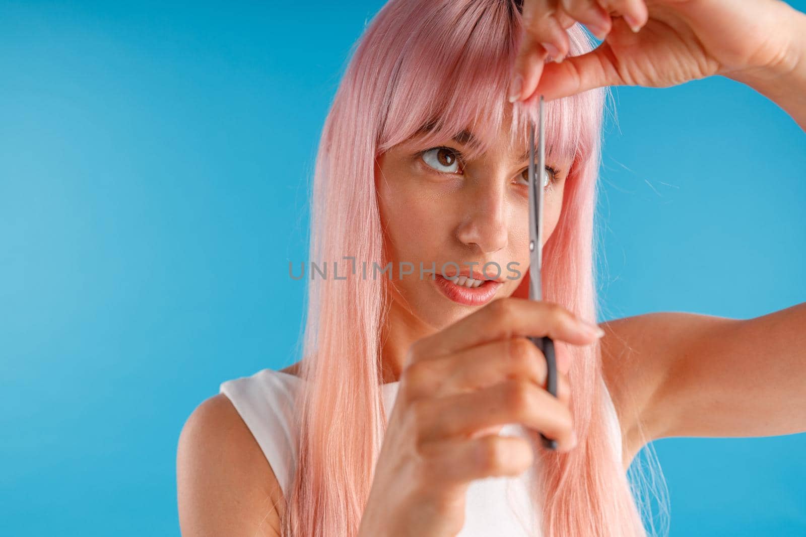 Portrait of cute young woman with pink hair cutting fringe herself using scissors, standing isolated over blue studio background. Beauty and hair care concept