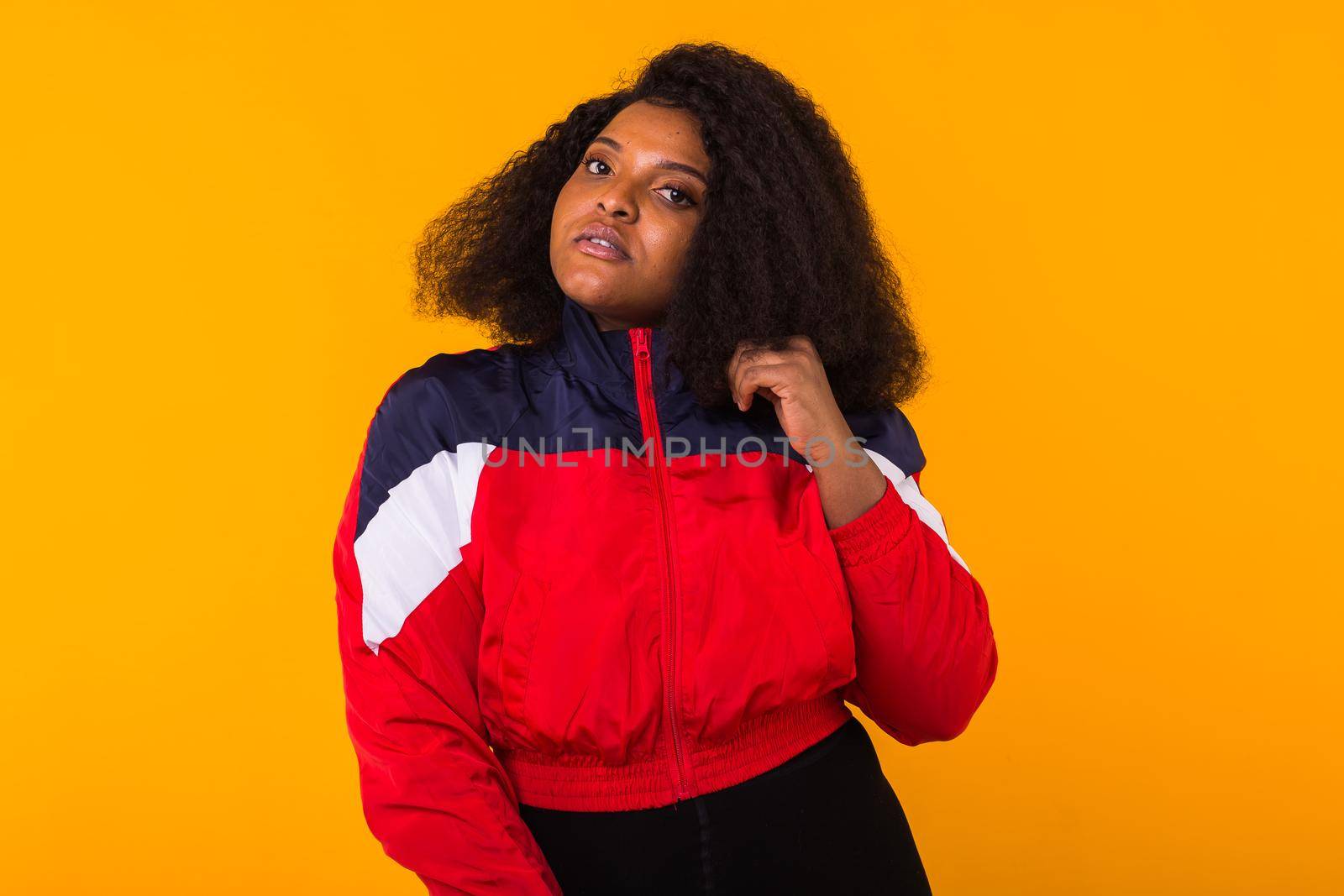 Funny curly african american girl dressed in the red sports jacket in the studio on yellow background. Fashion and people.