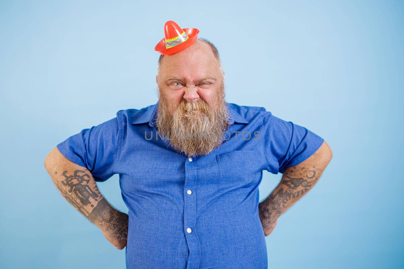 Funny bearded plus size man wearing small sombrero and tight blue shirt holds hands on waist grimacing on light blue background in studio