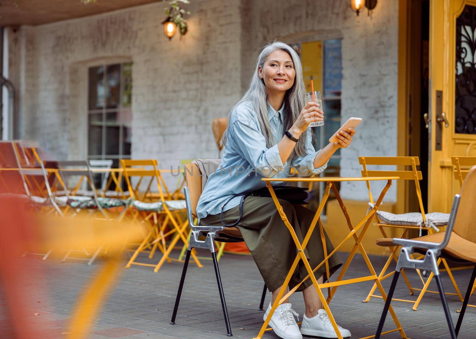 Elegant grey haired Asian woman with glass of water and smartphone sits at small table on outdoors cafe terrace