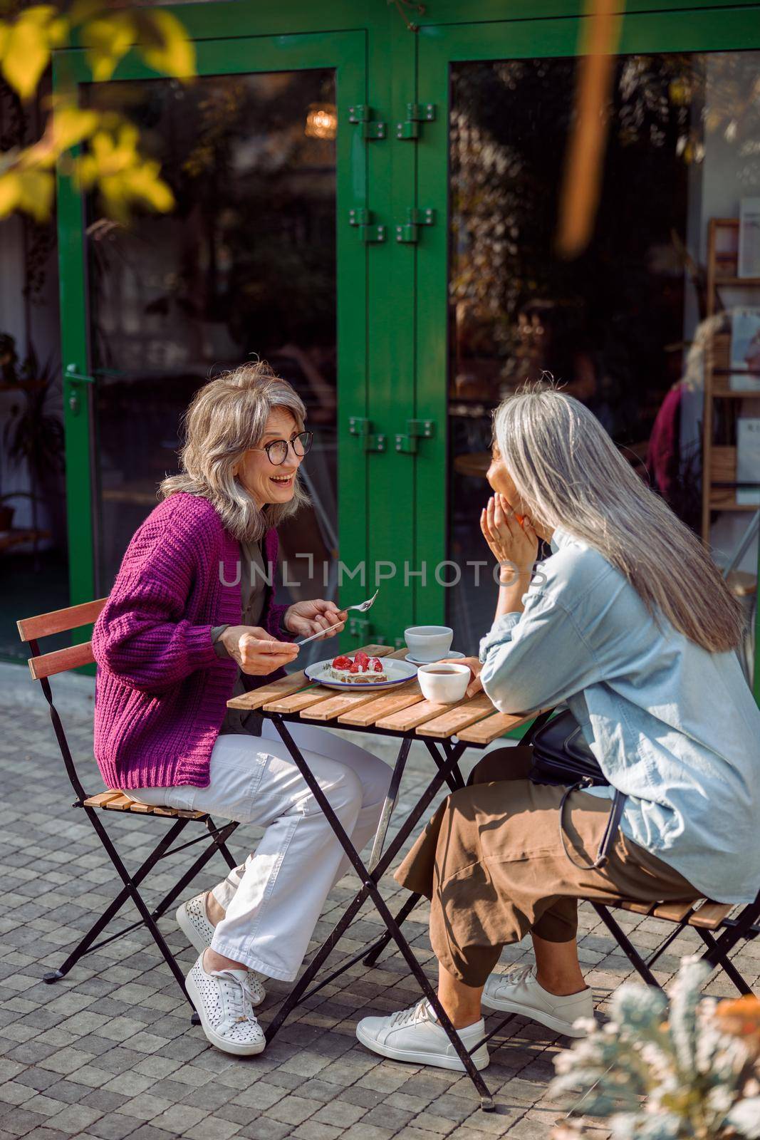 Joyful mature woman with long haired friend sit at small table with dessert and drinks on outdoors cafe terrace on autumn day