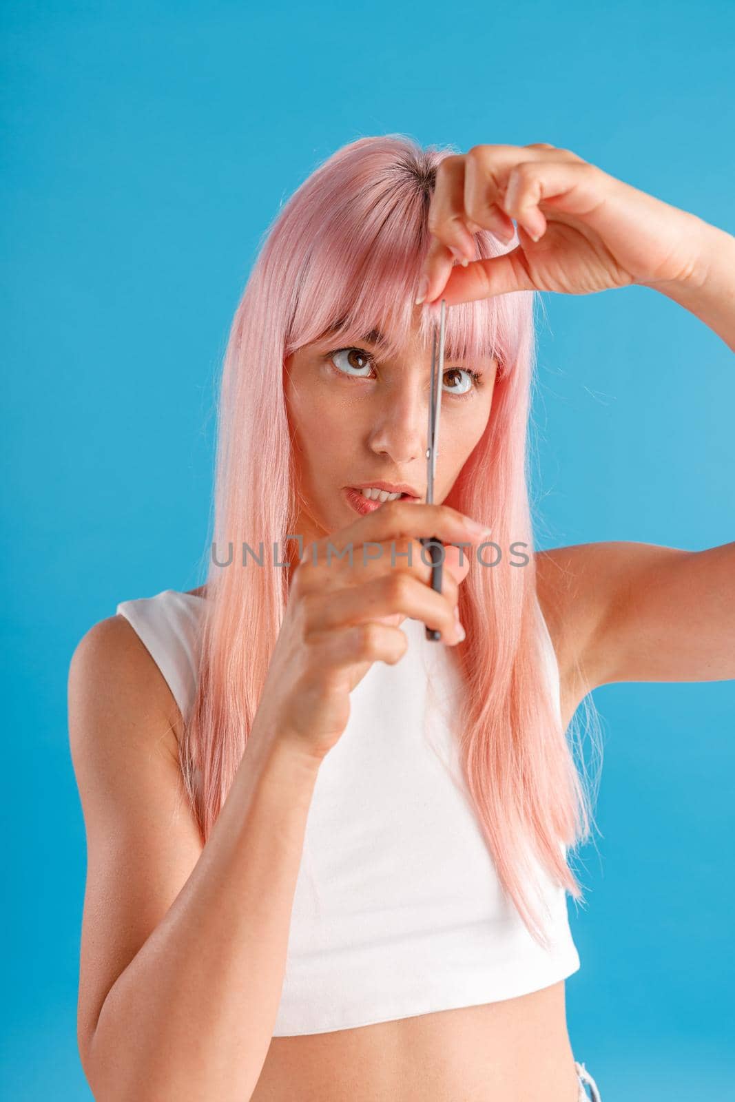 Cute young woman with pink hair cutting fringe herself using scissors, standing isolated over blue studio background. Beauty and hair care concept