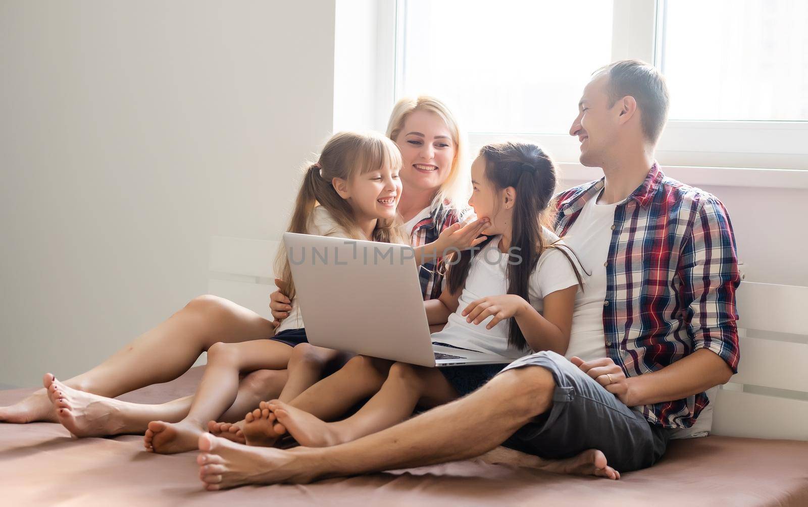 Happy family concept. Beautiful mother and handsome father with their daughters spending time together at home and lying on bed with laptop.