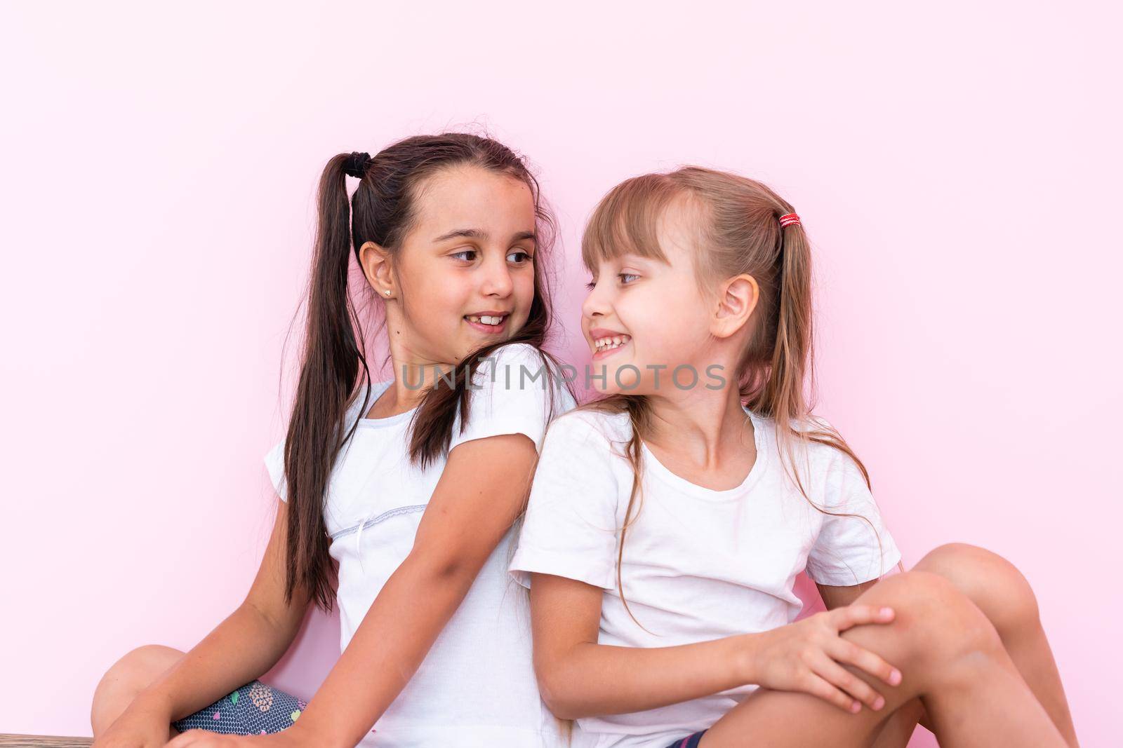 two little girl sitting on floor and looking up. Isolated on a pink background
