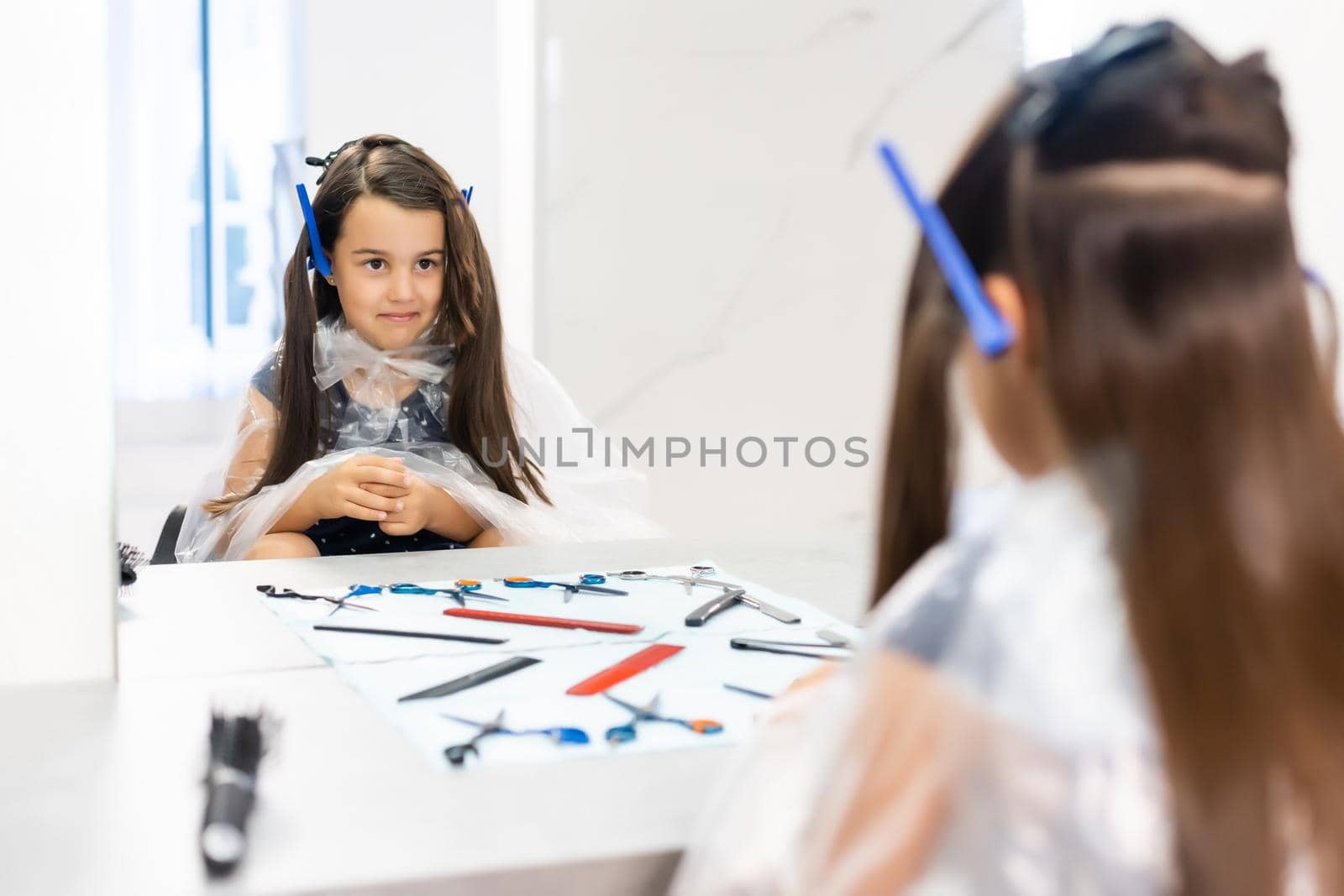Portrait of pretty little girl seating at table with accessories in a hairdressing salon
