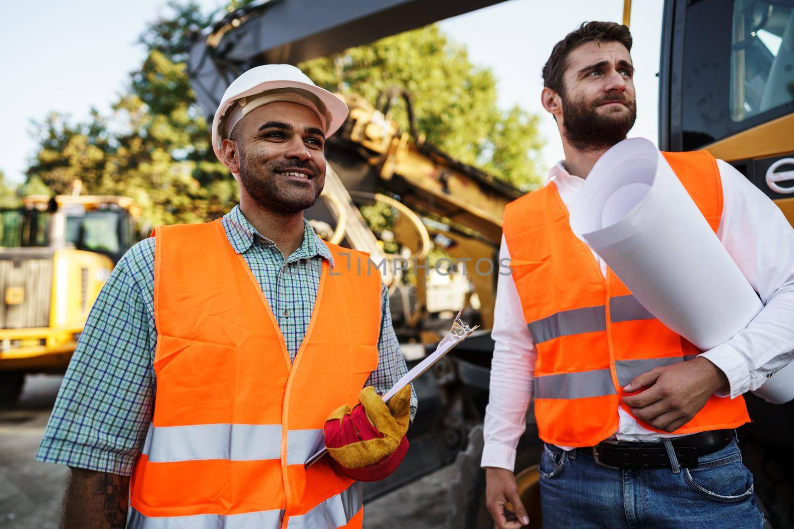 Two men engineers in workwear discussing their work standing against construction machines