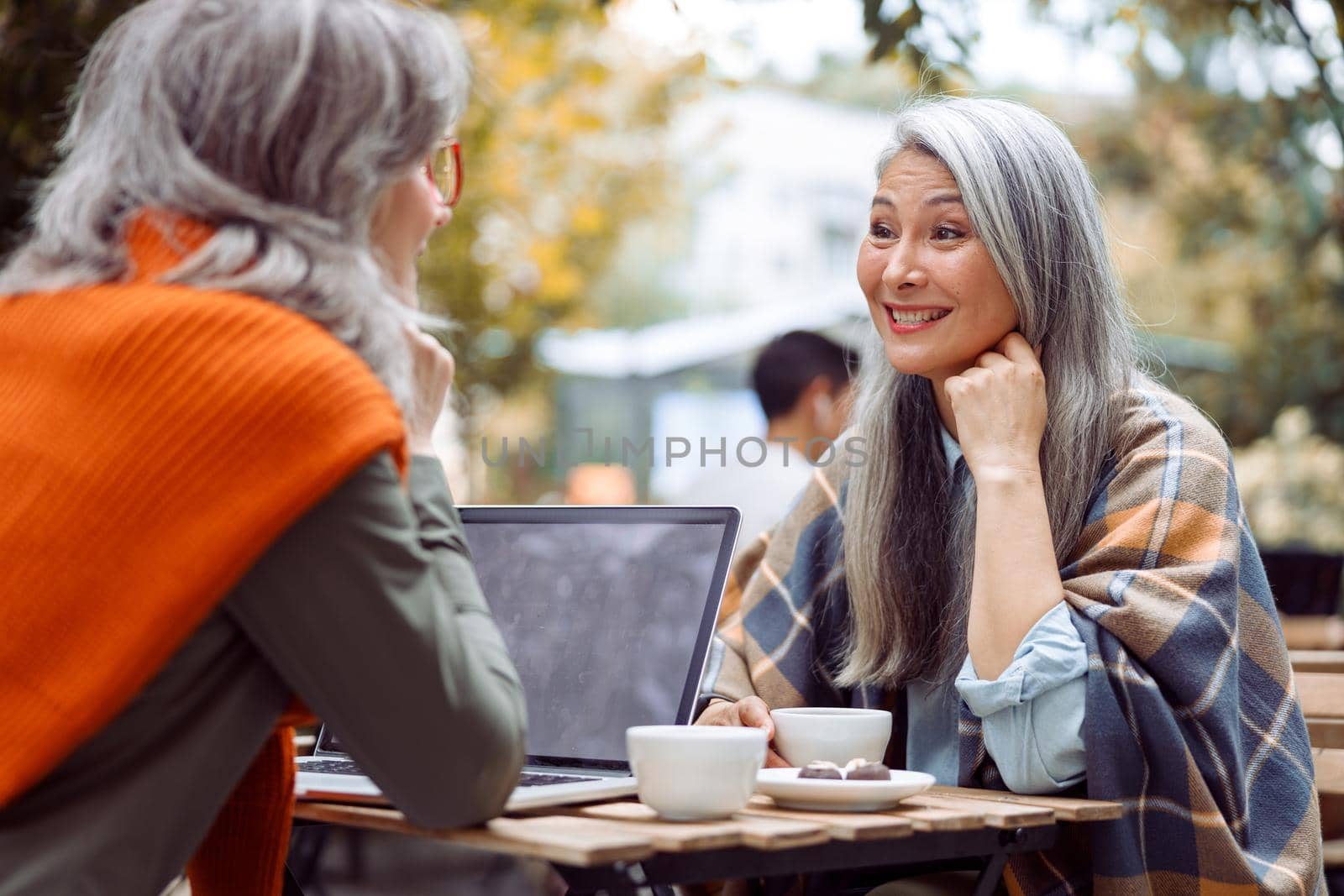 Positive mature Asian lady and grey haired friend with laptop sit together at table on outdoors cafe terrace cafe on nice autumn day