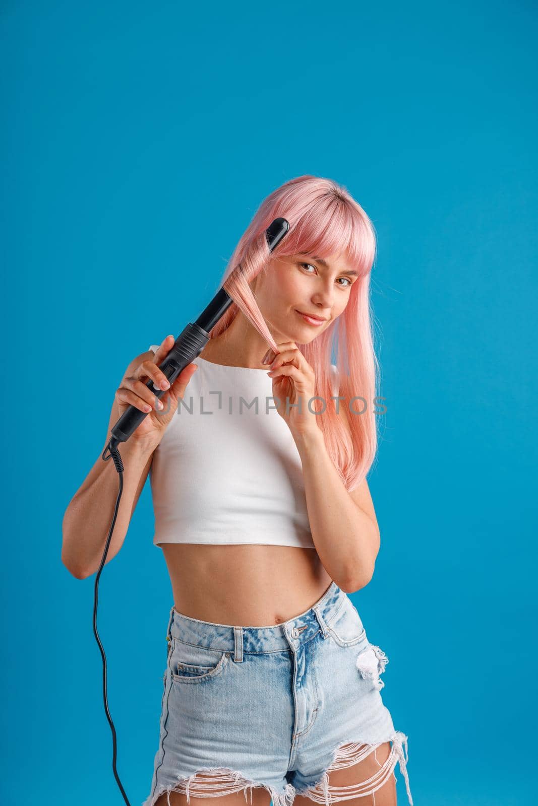 Attractive young woman looking at camera, curling pink long straight hair using curling iron, standing isolated over blue studio background. Beauty and hair care concept